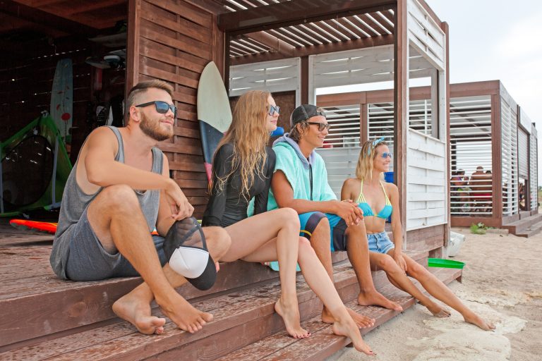 friendship, sea, summer vacation, water sport and people concept - group of friends wearing swimwear sitting with surfboards on beach
